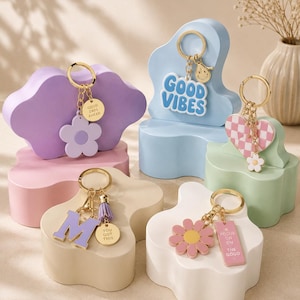 May include: A collection of keychains with gold-colored rings and charms. The charms include a purple flower, a blue cloud with "Good Vibes", a pink and white checkered heart, a purple letter "M", and a pink flower. Each keychain has a different charm with a positive message.