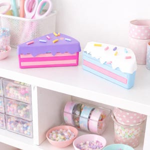 May include: A white shelf displays colorful stationery and decorative items. Two cake-shaped boxes with sprinkles sit on the top shelf. Below are small bowls of beads and containers of washi tape, all in pastel colors.