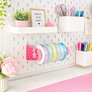 Puede incluir: Un organizador de pared de tablero perforado blanco con un dispensador de cintas rosa con patrón de panal que sostiene varios rollos de cinta pastel. Incluye estantes y recipientes blancos con bolígrafos, tijeras y un letrero que dice CREATE YOUR HAPPY.