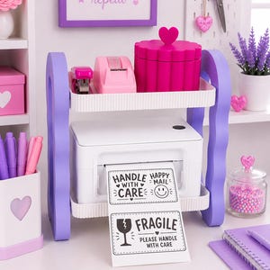 May include: A two-tiered purple and white desk organizer holding a white thermal label printer. The printer displays a label with the text HANDLE WITH CARE, HAPPY MAIL, and FRAGILE PLEASE HANDLE WITH CARE. The top shelf holds a pink stapler and box.