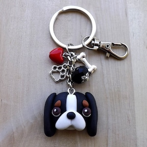 Puede incluir: Un llavero hecho a mano con un dije de cara de perro en blanco y negro, detalles marrones y una huella de pata plateada. Incluye un corazón rojo, una cuenta negra y un dije de hueso. El llavero está sujeto a un llavero y un cierre plateados.