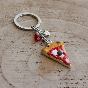 May include: A silver key chain with a red bead, a silver heart charm, and a pizza slice charm. The pizza slice is made of polymer clay and has red sauce, white cheese, and a green basil leaf.