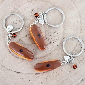 Könnte beinhalten: Drei Schlüsselanhänger mit Miniatur-Schokoladeneclairs mit einer Kaffeebohne oben drauf. Jeder Schlüsselanhänger hat einen silbernen Ring und eine silberne Kette mit einem Herz-Anhänger und braunen Perlen.