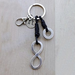 Pode incluir: Um chaveiro de prata com o símbolo do infinito com um cordão preto e dois anéis de prata. O chaveiro também tem um charme de prata com as letras "AL" nele.