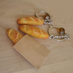 May include: A keychain featuring three miniature baguettes, one in a brown paper bag, with a silver chain and a brown bead.