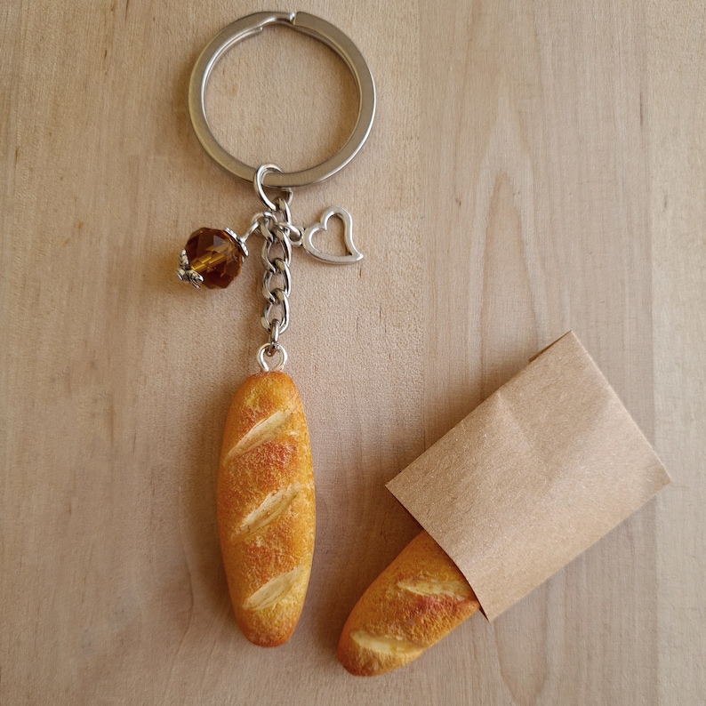 May include: A silver key chain with a brown crystal charm and a small heart charm. The keychain has a brown baguette charm attached to it. A brown paper bag with a brown baguette charm is also in the image.