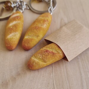 May include: Three small, golden brown, baguette-shaped keychains. One keychain is in a brown paper bag.