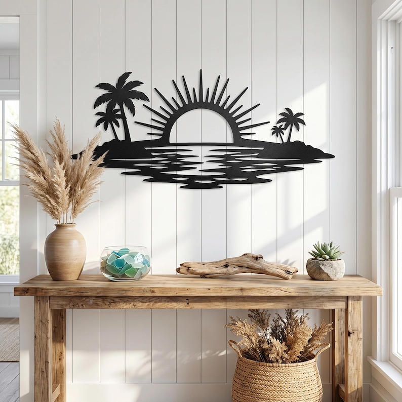 May include: A black metal wall art piece featuring a silhouette of a tropical island scene with palm trees, a rising sun with radiating beams, and a reflective water surface, mounted on a white vertical paneled wall above a wooden console table.