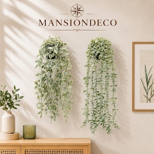May include: Two wall-mounted artificial hanging plants in black pots, featuring cascading green foliage with variegated leaves. Above the plants, the text MANSIONDECO is displayed with a compass icon. The plants are positioned on a light wall.