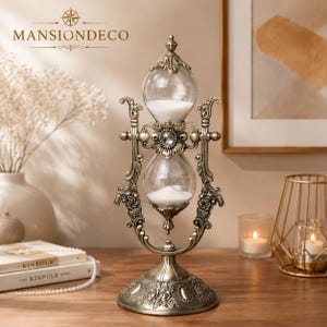 May include: An antique-style brass hourglass featuring a rotating frame with intricate floral engravings and a central crystal accent. The two glass bulbs contain white sand, supported by a decorative circular base with detailed metalwork.