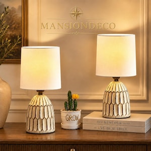 May include: Two table lamps with off-white cylindrical fabric shades and textured, cream-colored ceramic bases featuring a repeating geometric honeycomb pattern. The lamps emit a warm glow and are positioned on a wooden surface with a small plant.