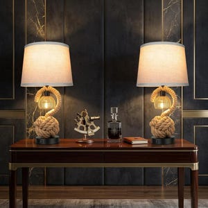 May include: Two table lamps with beige fabric drum shades, rope-wrapped bases featuring a large knot design, and a small woven cage light element. Between the lamps sits a brass nautical sextant, a glass decanter, and a small leather-bound book.