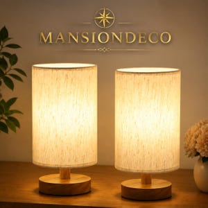 May include: Two cylindrical table lamps with light-colored fabric shades and circular wooden bases. The lamps emit a warm glow. Above the lamps, the text MANSIONDECO is displayed in gold lettering with a compass rose icon centered above the brand name.