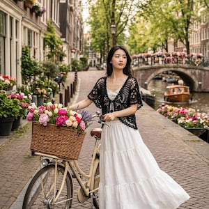 May include: A woman in a white dress and black crochet top stands next to a cream-colored bicycle with a wicker basket filled with flowers. The scene is set on a brick-paved street in a European city with a canal and bridge in the background.