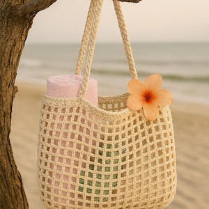 May include: A beige crocheted tote bag with rope handles hangs from a tree branch. The bag contains a rolled-up pink towel and a green item. An orange flower is attached to the side of the bag. The background is a sandy beach.