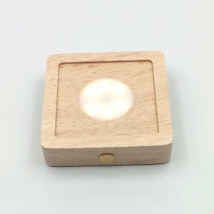 Könnte beinhalten: Ein quadratischer Holzsockel mit integrierter LED-Leuchte. Das Licht ist ein heller, leuchtender Kreis in der Mitte des Holzes. Auf der Vorderseite des Sockels befindet sich ein kleiner, runder, goldfarbener Knopf. Das Holz hat eine natürliche, helle Farbe.
