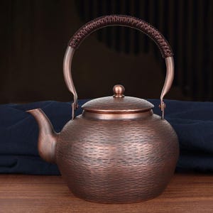 May include: A copper-colored teapot with a textured surface and a woven handle. The teapot has a curved spout and a lid with a small knob. The handle is brown and arched. The teapot is designed for brewing tea.