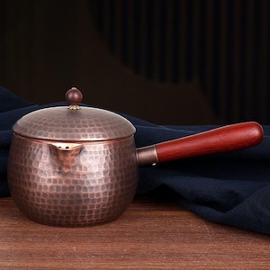 Puede incluir: Tetera de color cobre con textura martillada, asa de madera y tapa a juego. La tetera tiene un pequeño pico y un pomo de madera en la parte superior de la tapa. El asa es de color marrón oscuro.