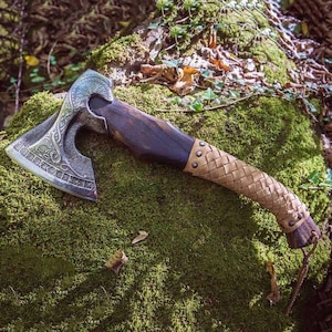 May include: A detailed axe with a silver-colored head featuring intricate carvings and a dark wooden handle. The handle transitions to a woven leather grip, secured with rivets. The axe rests on a bed of green moss, suggesting an outdoor setting.