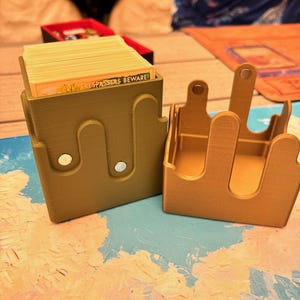 May include: A 3D-printed olive green card deck box with a wavy top edge and magnetic closures, containing a stack of cards with the visible text TRESPASSERS BEWARE! next to a matching empty box lid with three upward-extending tabs and magnets.