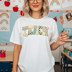 Op de afbeelding: Wit T-shirt met het woord "TEACH" in een patchwork-ontwerp. De letters zijn samengesteld uit verschillende patronen, waaronder bloemen, ruiten en stippen. Een madeliefje staat aan de linkerkant van het woord.