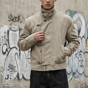 May include: A beige cotton jacket featuring a high stand-up collar with snap buttons, a front panel secured by three metal toggle clasps, long sleeves with cuffs, and a straight hemline. The garment is worn over dark denim trousers.