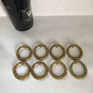 Könnte beinhalten: Acht antike Messing-Ringgriffe mit strukturiertem Design sind auf einer weißen Oberfläche angeordnet. Im Hintergrund befindet sich eine dunkle Flasche. Diese dekorativen Griffe sind wahrscheinlich für Möbel.