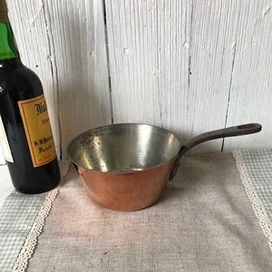 May include: A copper saucepan with a dark brown wooden handle. The saucepan is sitting on a white and gray checkered tablecloth.