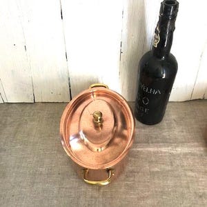 Puede incluir: Una olla de color cobre con tapa y asas doradas, y una botella negra con etiqueta. La olla es redonda con tapa y dos asas. La botella es oscura y tiene un cuello largo.