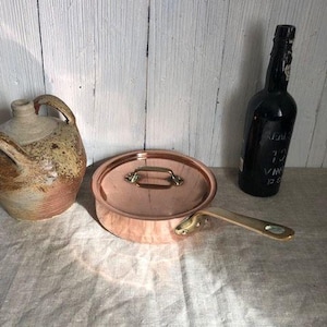 May include: A copper saucepan with a lid and a wooden handle. The saucepan is sitting on a white linen tablecloth.