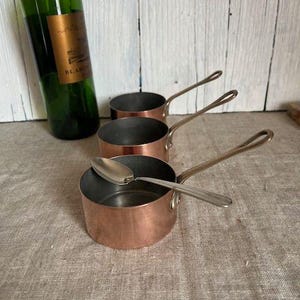 May include: Three small copper saucepans with long handles, one with a silver spoon inside. A green wine bottle with a gold label is in the background. The pans are on a neutral-colored linen surface.