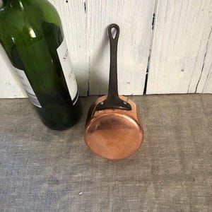 Può includere: Una piccola casseruola di rame con un lungo manico scuro. La pentola ha una base arrotondata e una superficie lucida e riflettente. Accanto alla casseruola c'è una bottiglia di vino verde con tappo rosso. Sfondo bianco in legno.