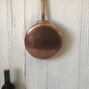 May include: A copper frying pan with a brass handle hanging on a metal rack against a white wood background.