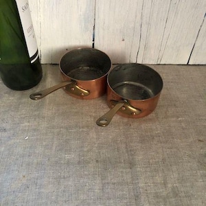 May include: Two small copper saucepans with brass handles sit on a gray linen surface. The pans have a shiny copper exterior and a dark interior. A green wine bottle with a white label is partially visible in the background.