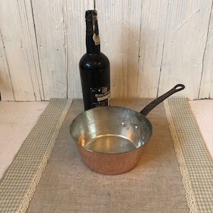 May include: A copper saucepan with a black handle sits on a beige and white checkered tablecloth. A dark glass bottle with a label that reads "Royal Crown" is in the background.