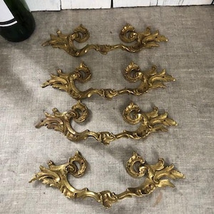 Könnte beinhalten: Vier antike Schubladengriffe aus Messing mit einem floralen, geschwungenen Design. Die Griffe liegen auf einer grauen Leinenoberfläche, mit einer grünen Flasche im Hintergrund. Geeignet für Möbelrestaurierung.