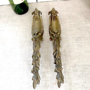 Op de afbeelding: Twee antieke, bronskleurige decoratieve metalen applicaties met sierlijke blad- en bloemmotieven. De applicaties zijn verticaal georiënteerd en hebben een gedetailleerd, getextureerd oppervlak. Een groene wijnfles is zichtbaar op de achtergrond.