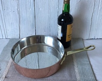 May include: A copper-bottomed, stainless steel frying pan with a brass handle sits on a gray and white checkered tablecloth. A dark glass bottle with a green and gold label is behind the pan.