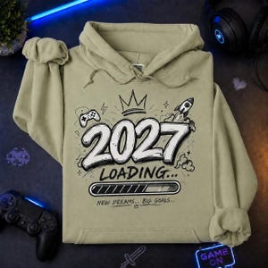 May include: A light olive green hooded sweatshirt featuring a graphic print with the text 2027 LOADING... NEW DREAMS... BIG GOALS... along with illustrations of a game controller, a crown, a rocket ship, and a progress loading bar.