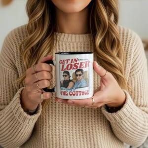 May include: White ceramic mug with a black interior and handle. The mug features the text "GET IN LOSER" above an illustration of two people in a car, with the text "We're going to THE COTTAGE" below. The mug is held by a person.