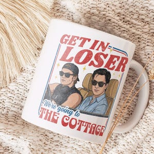 Könnte beinhalten: Weiße Keramik-Tasse mit dem Text "GET IN LOSER" in Rot und Blau sowie einer Illustration von zwei Personen in einem Auto. Die Tasse trägt auch den Text "We're going to THE COTTAGE".