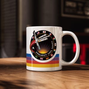 Puede incluir: Taza de cerámica blanca con el logo "Project Hail Mary". El diseño incluye un emblema circular con un cohete, banderas y el texto "Project Hail Mary". La taza tiene un diseño de rayas de arcoíris en la parte inferior.