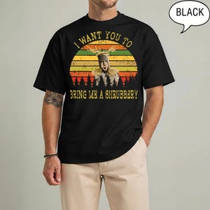 May include: A black short-sleeve t-shirt featuring a graphic of a knight in a helmet with antlers. The design includes a sunset background, a forest silhouette, and the text: I WANT YOU TO BRING ME A SHRUBBERY. A speech bubble says BLACK.