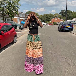 May include: A black sleeveless mock neck top paired with a long, tiered maxi skirt featuring horizontal bands of green, tan, orange, dark purple, and pink, each decorated with a distinct white patterned print and an adjustable drawstring waist.