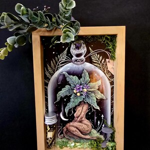 May include: A framed artwork featuring a woman-like figure under a glass dome, adorned with a plant with purple flowers. A small vial with a silver cap and a miniature mushroom table are also present. The artwork is set against a dark background.