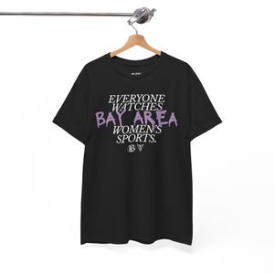 以下が含まれることがあります： 白と紫で「EVERYONE WATCHES BAY AREA WOMEN'S SPORTS」と書かれた黒いTシャツ。木製のハンガーにかかっています。