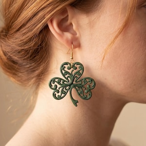 May include: Green shamrock earrings with intricate filigree detailing. The earrings have a gold-colored hook and are worn on a person's ear. The earrings are a St. Patrick's Day theme.