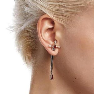 Könnte beinhalten: Ein silberfarbener Ohrring mit einem kreuzförmigen Stecker, der einen zentralen roten Stein und klare Akzente aufweist. Ein langer, schlanker silberfarbener Dorn hängt vertikal am Stecker und endet in einem kleinen, tropfenförmigen roten Stein.