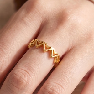 Puede incluir: Un anillo dorado con un diseño de corazones repetidos. El anillo se lleva en un dedo, mostrando el intrincado patrón de corazón abierto. El tono cálido y el diseño delicado del anillo lo convierten en un accesorio elegante.