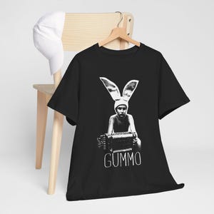 Puede incluir: Una camiseta negra de manga corta con un estampado gráfico blanco de una persona con orejas de conejo que sostiene un acordeón, con el texto GUMMO impreso debajo. Una gorra de béisbol blanca lisa cuelga del respaldo de la silla de madera.